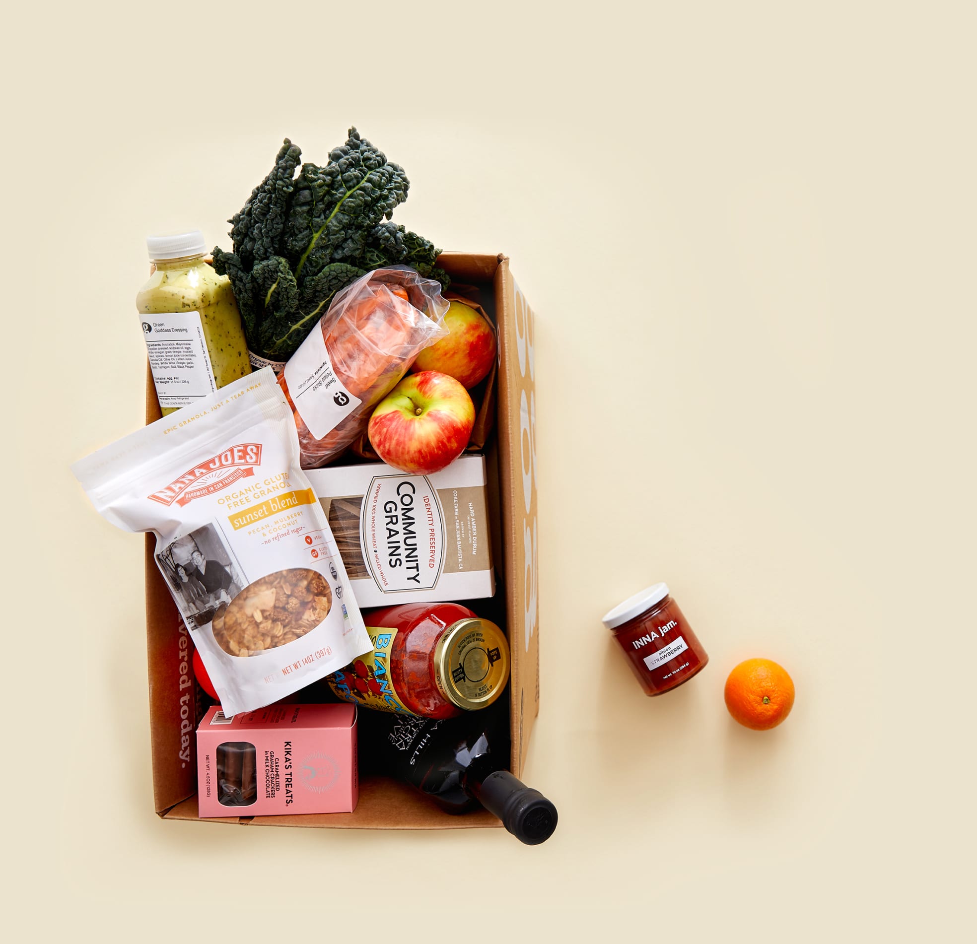 Bird's-eye view of a Good Eggs cardboard box of groceries including fresh vegetables, fruit, juice and jam on a beige surface.